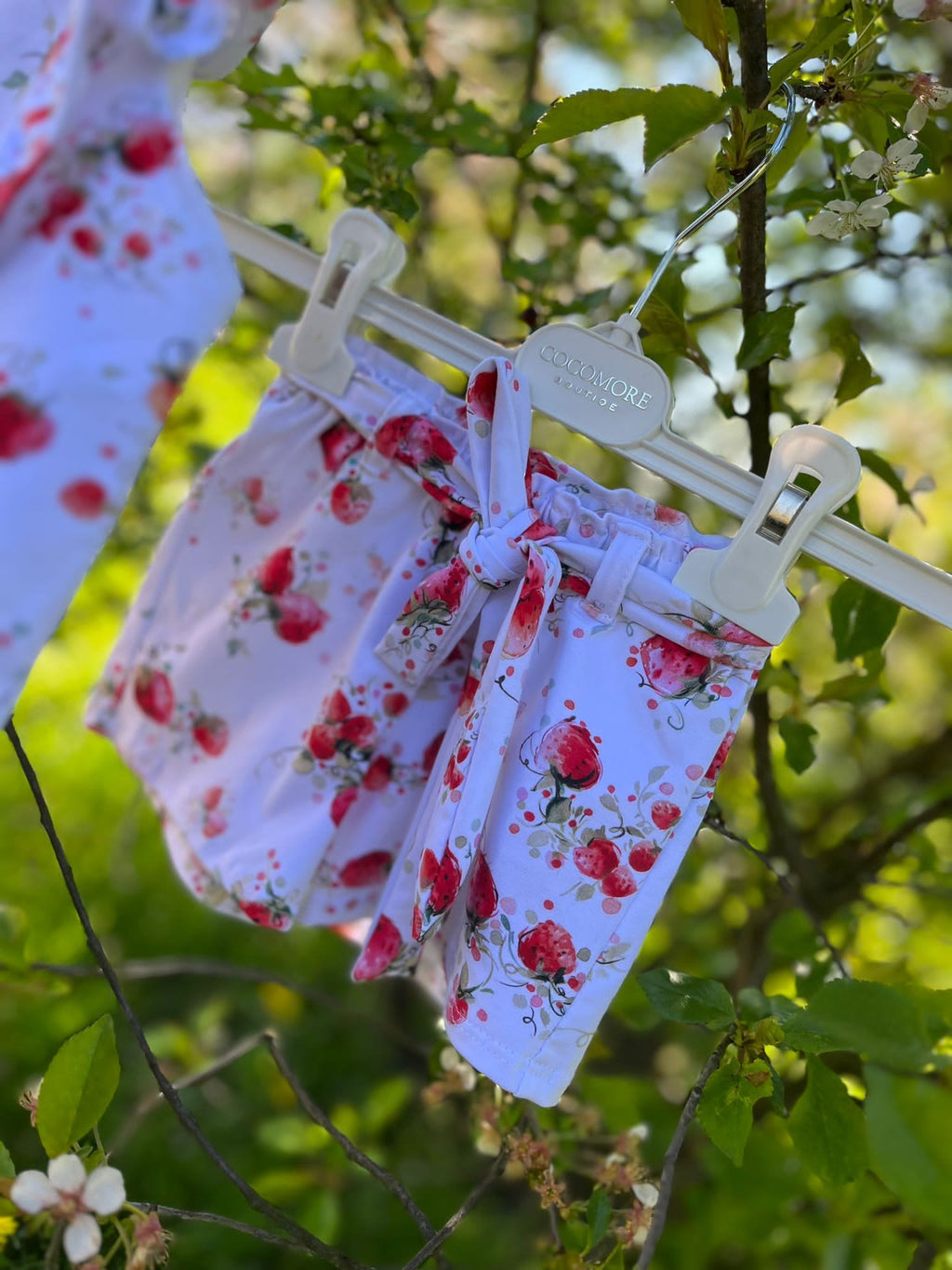 Witte meisjes short met aardbeienprint en strikdetail, gefotografeerd buiten tussen groene bladeren.
